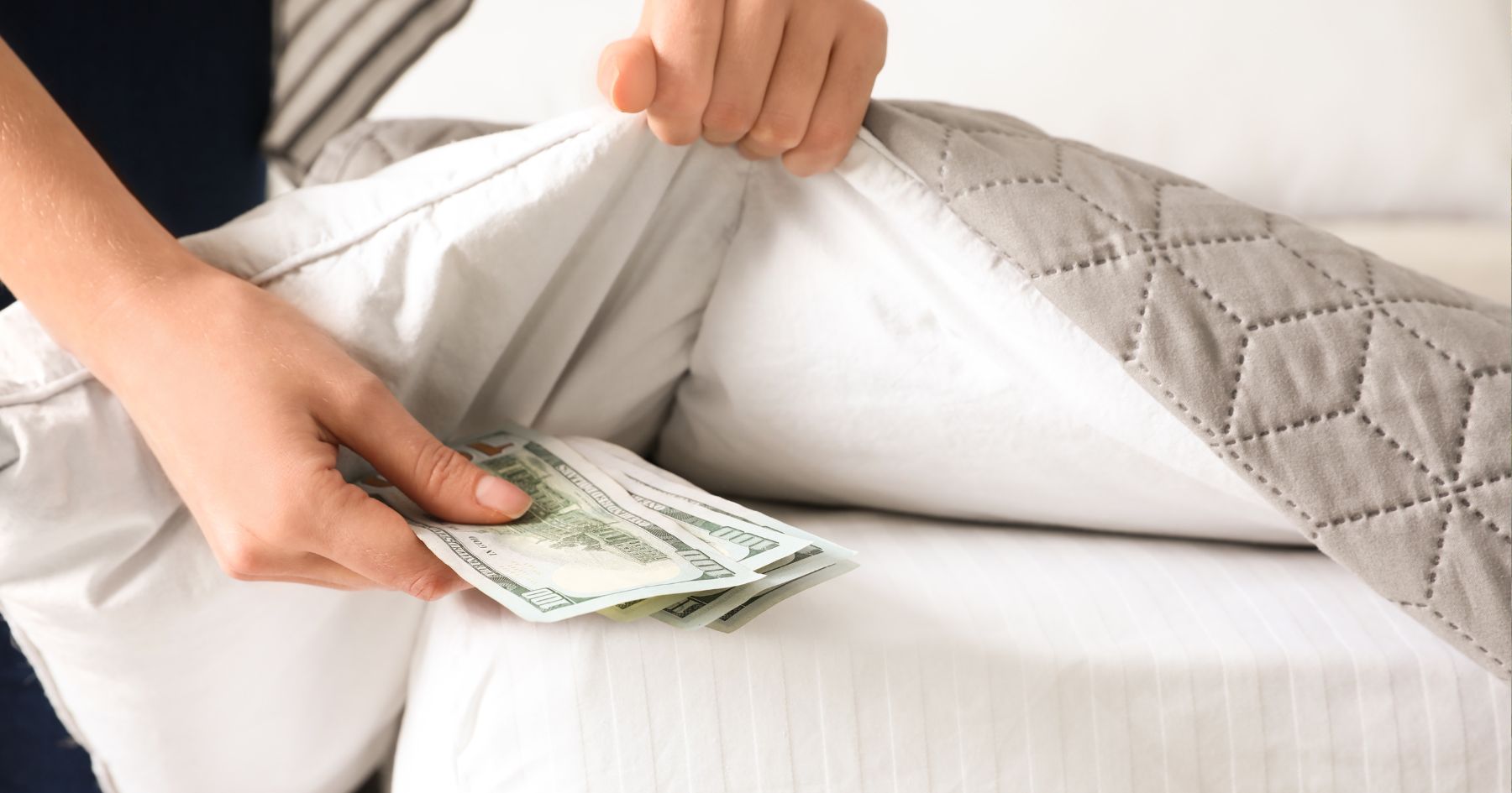 A person hides dollar bills under a mattress in a room with white bedding and a gray quilted bed cover.