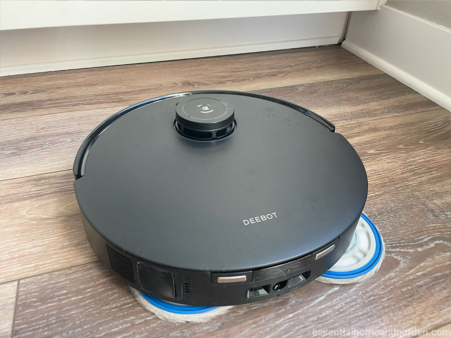 A black DEEBOT t30s robot vacuum cleaner mopping a wooden floor near a white baseboard.